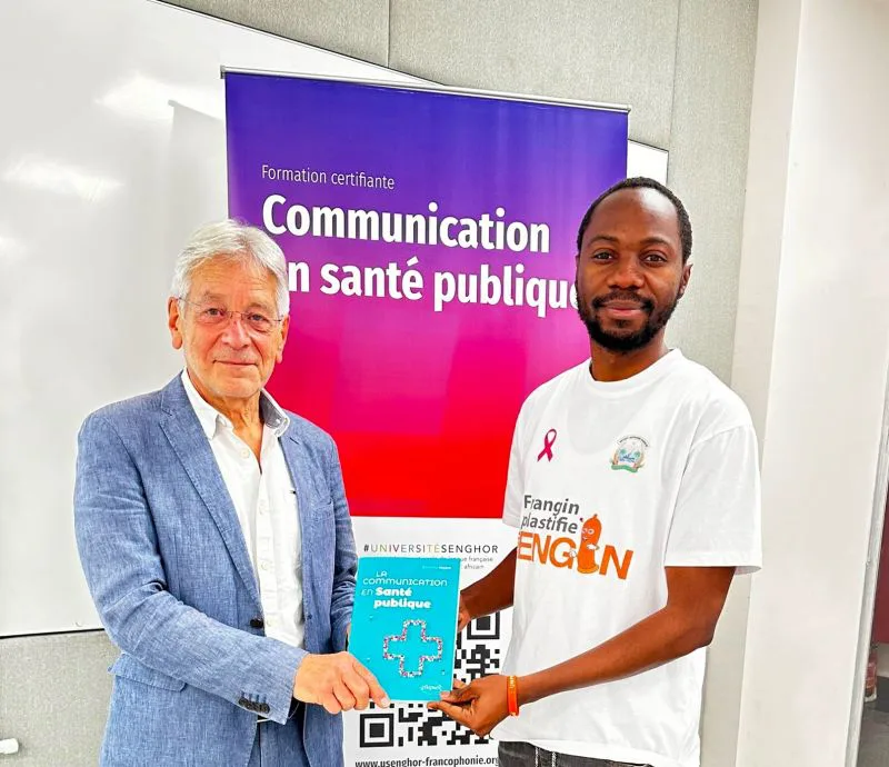 Bernard Seytre remet un exemplaire dédicacé de La communication en santé publique à Aimé Batuyingila, devant l’affiche de la formation certifiante de l’université Senghor